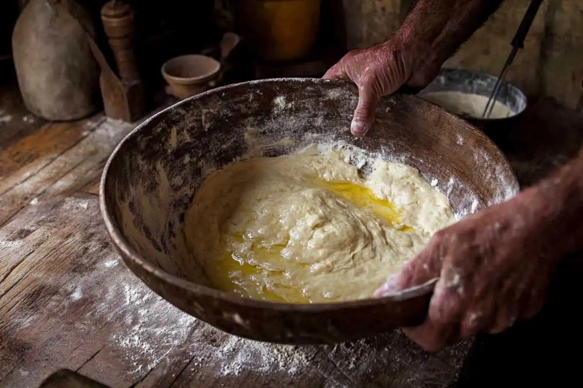 Mixing-Sourdough-Dough-in-a-Rustic-Wooden-Bowl A close-up of hands mixing sourdough dough in a rustic wooden bowl, with a drizzle of oil on the surface.