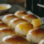 A pastry brush applying egg wash to risen sourdough brioche buns on a baking sheet.