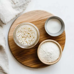 A flat-lay of sourdough bread ingredients: starter, flour, salt, and water on a wooden board.
