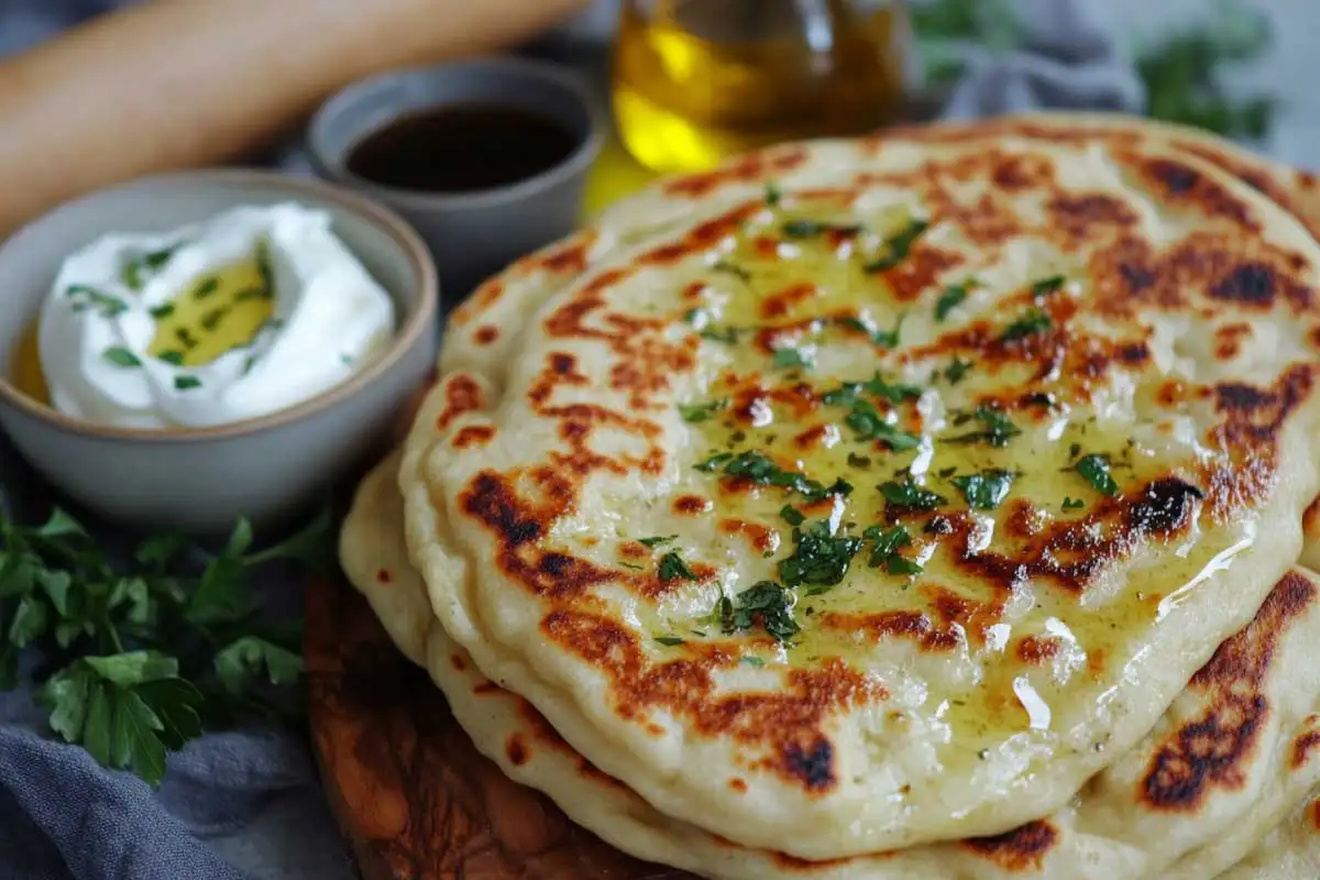 A stack of golden sourdough naan brushed with ghee, garnished with herbs, and served with yogurt and oil.